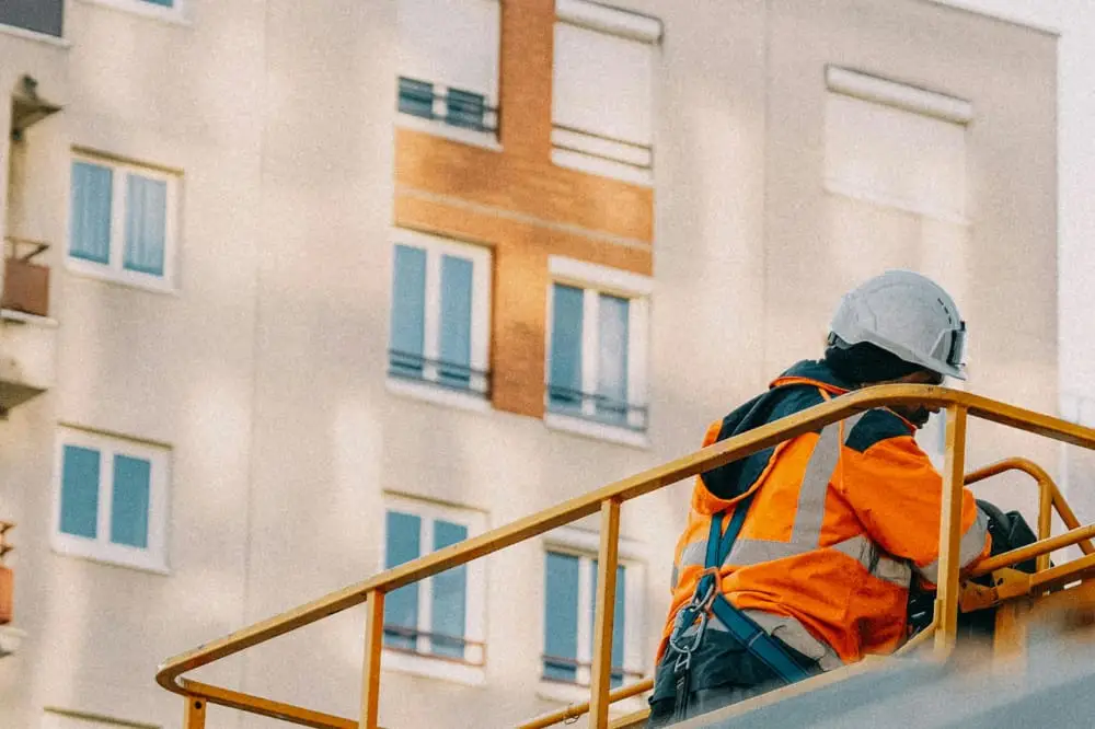 A construction worker on some kind of platform with a railing, with a building in the background